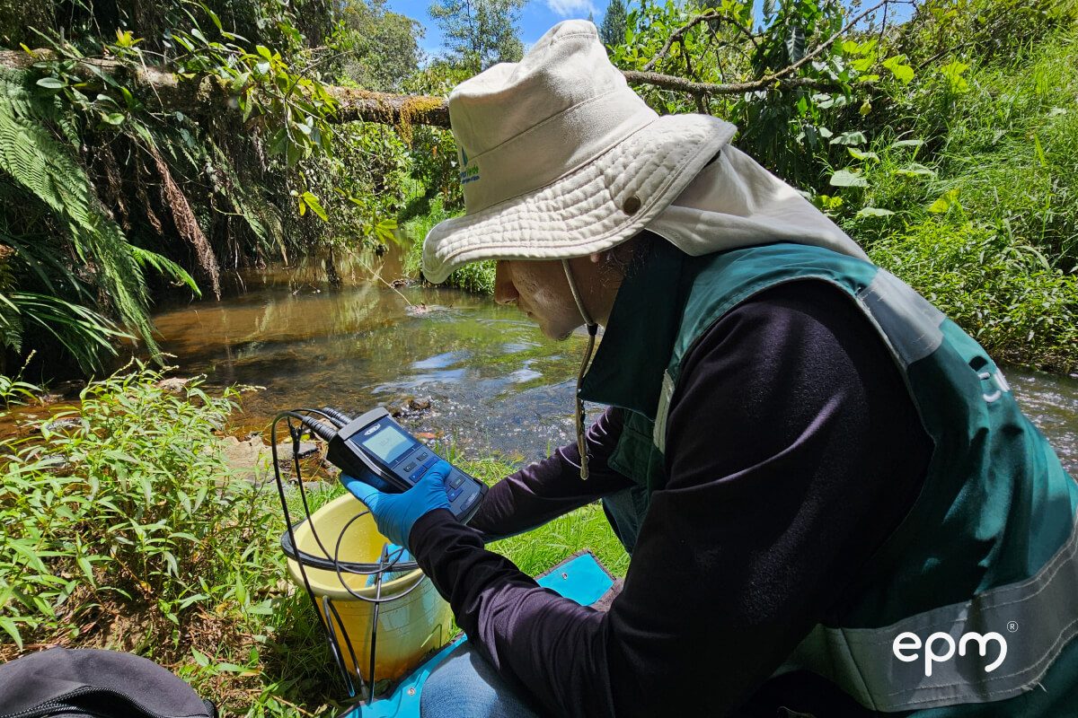 Fotografía de un hombre con sombrero de ala ancha y guantes azules realiza pruebas de calidad del agua en un riachuelo rodeado de vegetación.