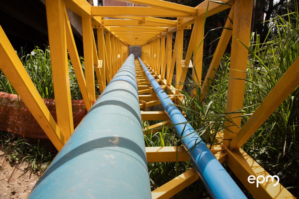 Vista en perspectiva de una pasarela de soporte amarillo con dos grandes tuberías azul y celeste pasando por el centro, rodeada de vegetación.