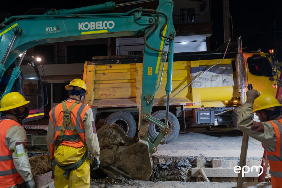 Trabajadores de EPM con cascos y chalecos naranjas dirigiendo una excavación nocturna.