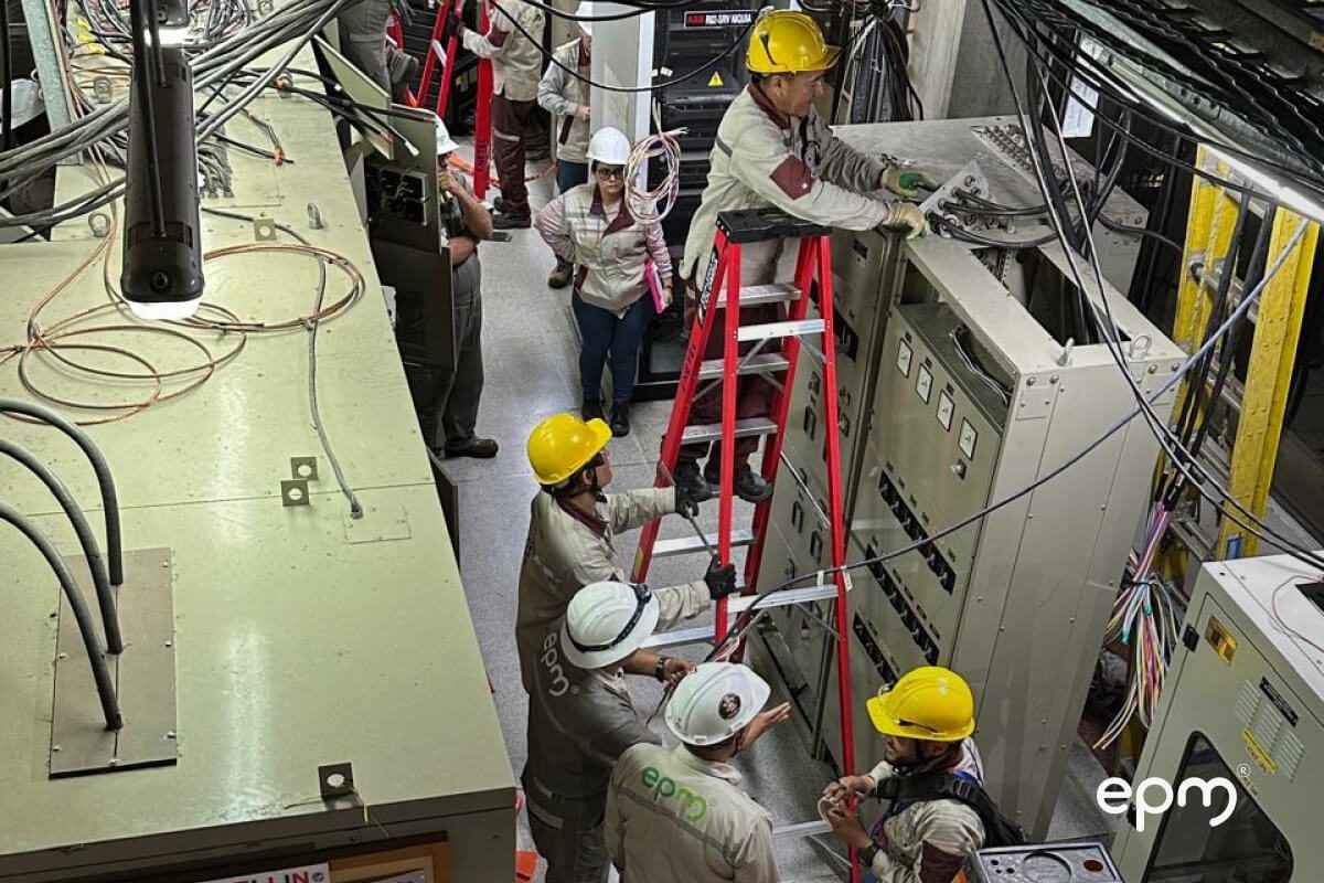 Fotografía de un grupo de trabajadores con cascos amarillos y ropa de trabajo están de pie alrededor de maquinaria y cables, uno de ellos está encima de unas escaleras rojas, trabajando en un panel de control. 