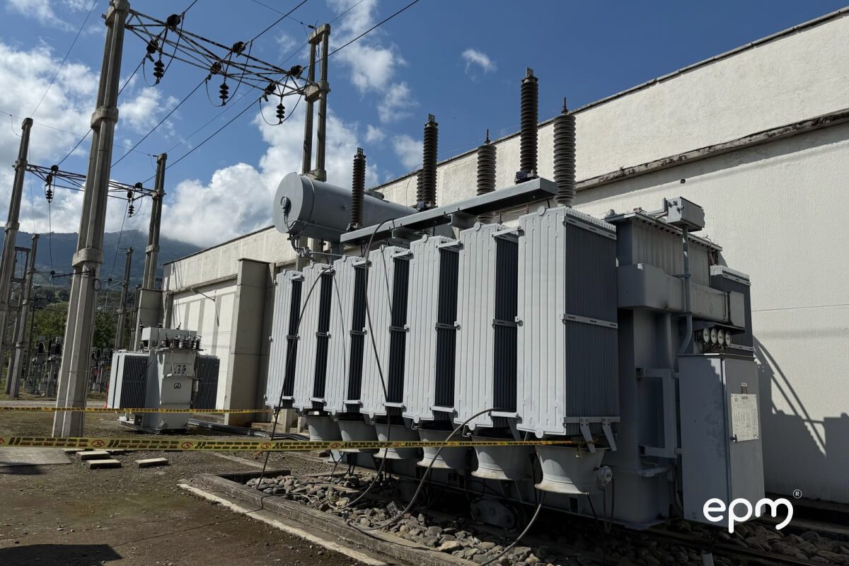 Fotografía de un transformador eléctrico gris grande con aisladores de porcelana, ubicado en una subestación al aire libre con cables y otros equipos eléctricos.