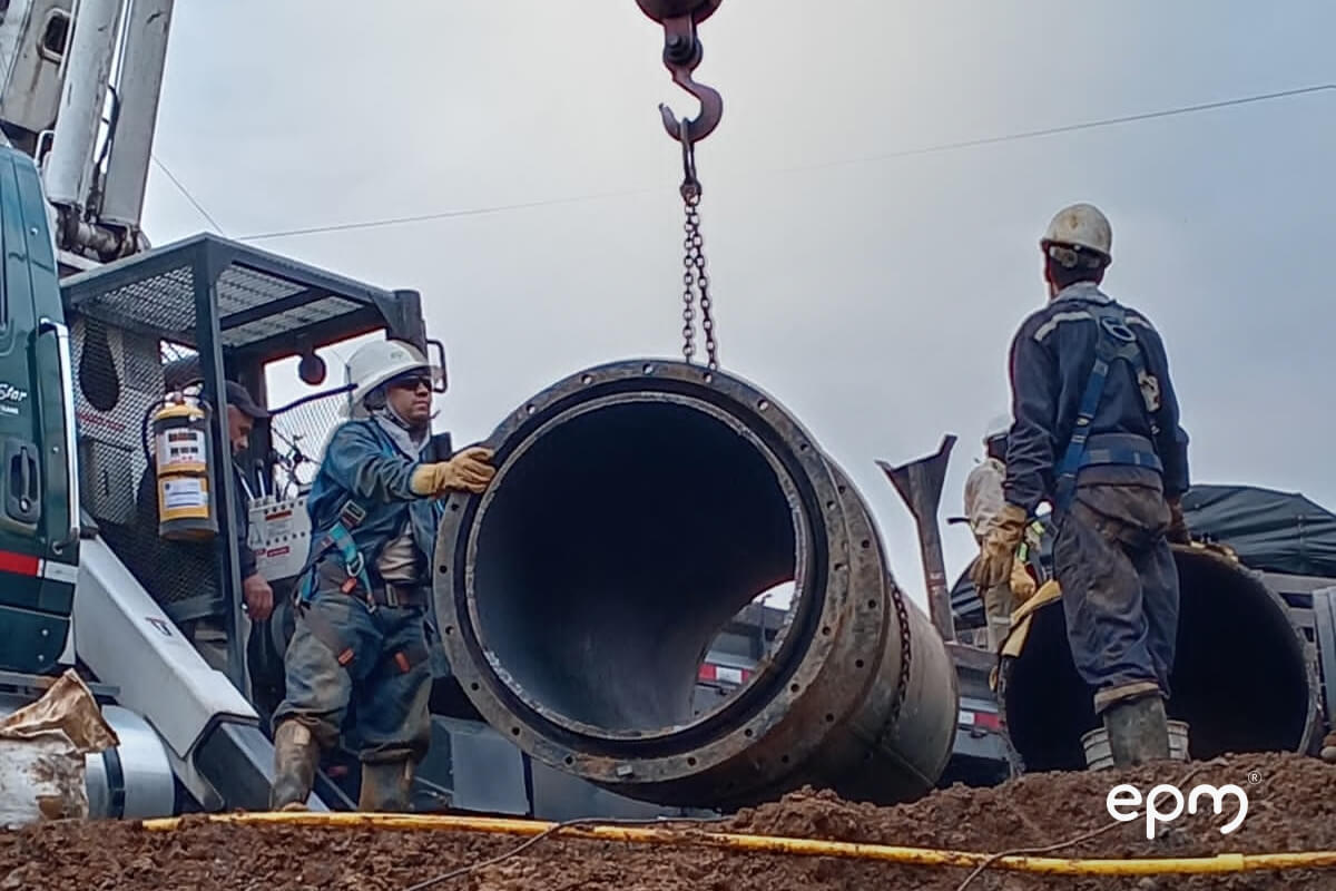 Trabajadores de EPM con cascos y arneses de seguridad guían una gran sección de tubería metálica suspendida por una grúa, en un área de obra sobre tierra.