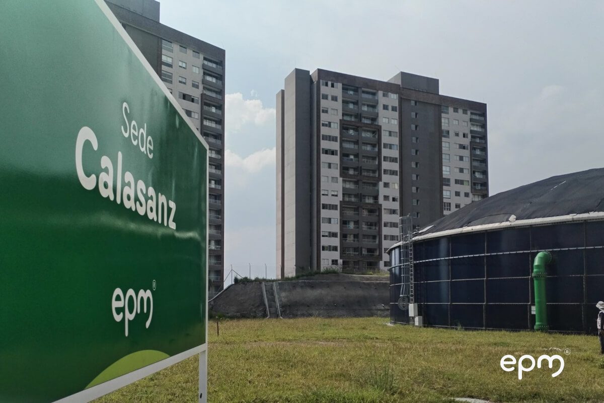 Fotografía de Vista de la sede Calasanz de EPM, con un letrero verde en primer plano y edificios residenciales altos al fondo, junto a una estructura de almacenamiento de agua.