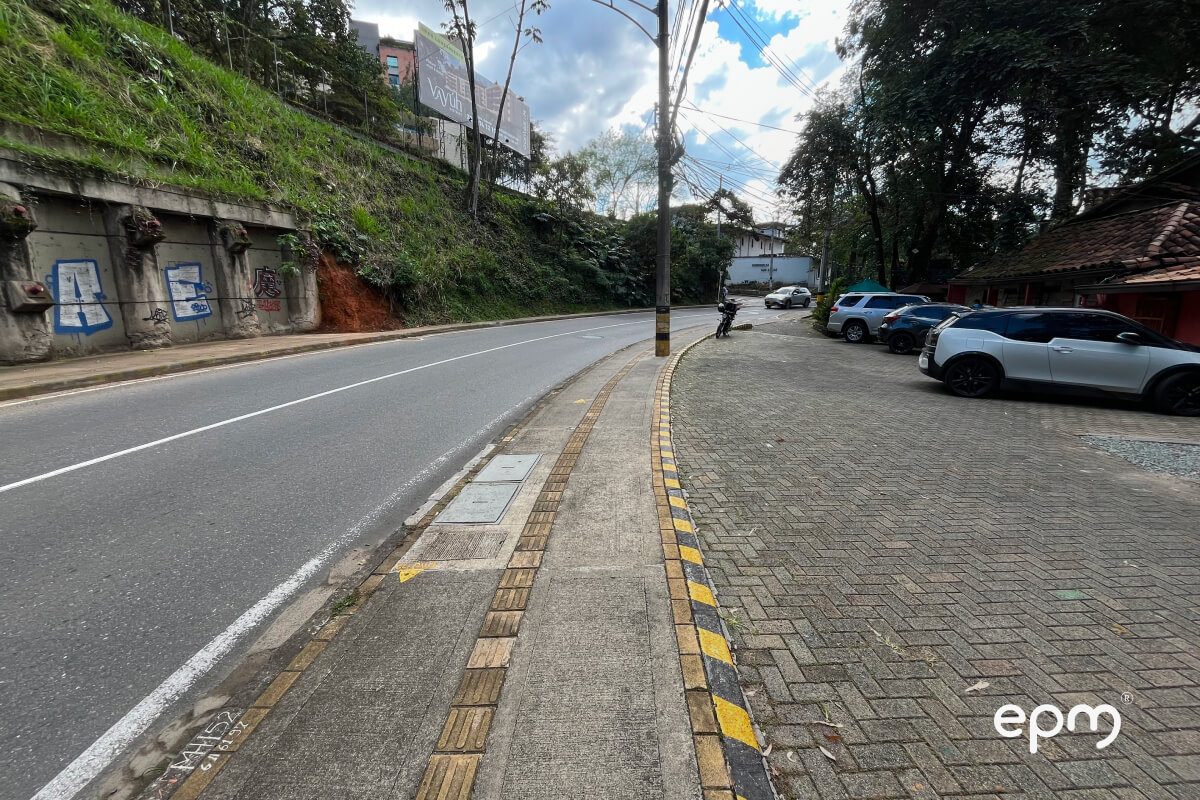 Fotografía de calle en un área urbana con ciclovía y andén delimitados por líneas amarillas y negras, rodeada de vegetación, con algunos vehículos estacionados y una casa con techo de tejas al fondo.