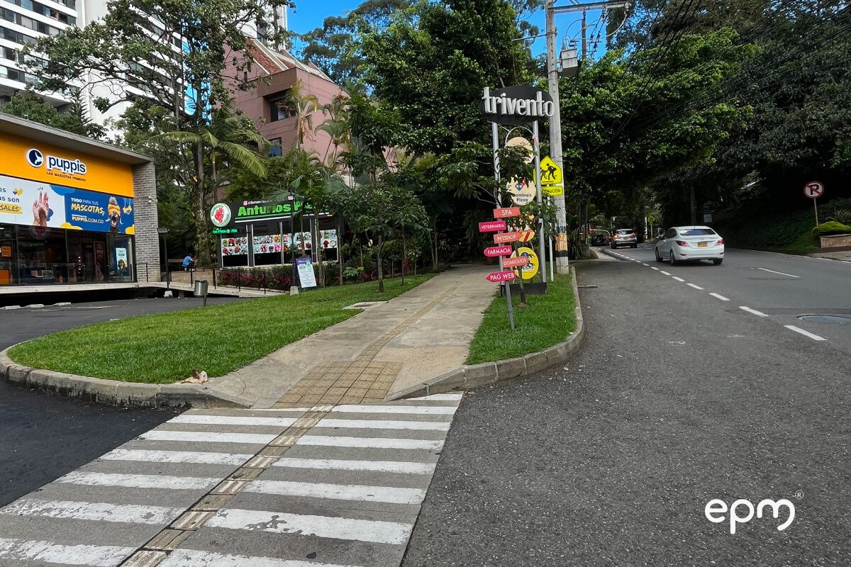 Fotografía de una calle urbana con camino peatonal, letreros de tiendas y tráfico vehicular