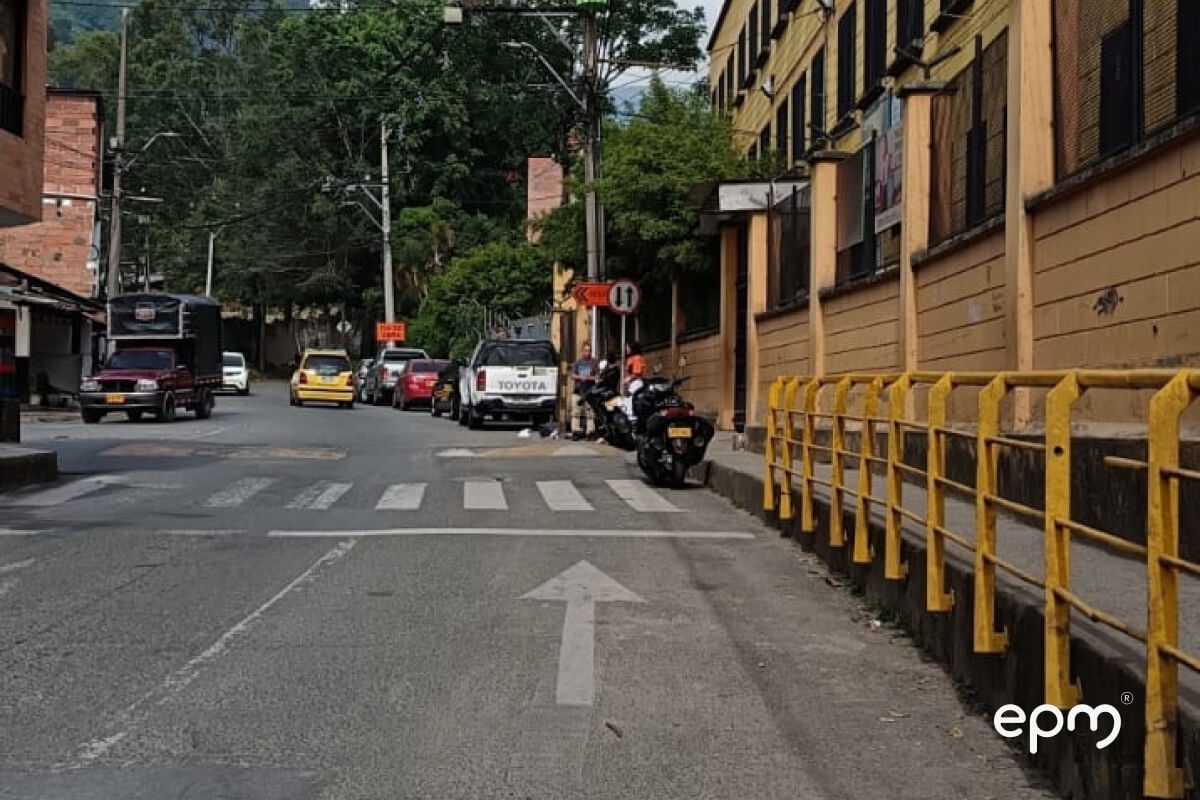 Fotografía de una calle urbana con varios vehiculos estacionados, la calle tiene una flecha señalando un cruce de peatones y barandilla amarilla del lado derecho
