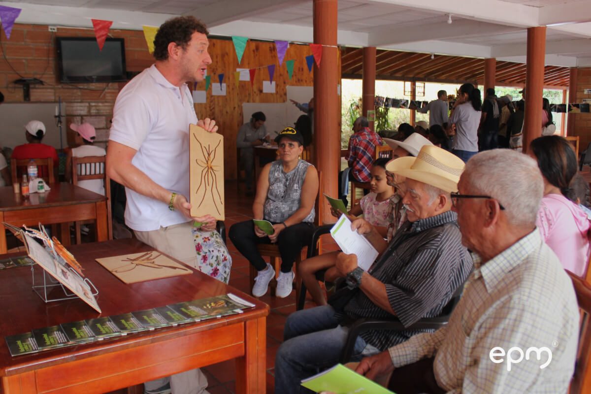Fotografía de taller educativo en un espacio comunitario donde un hombre sostiene una tabla con la ilustración de un insecto