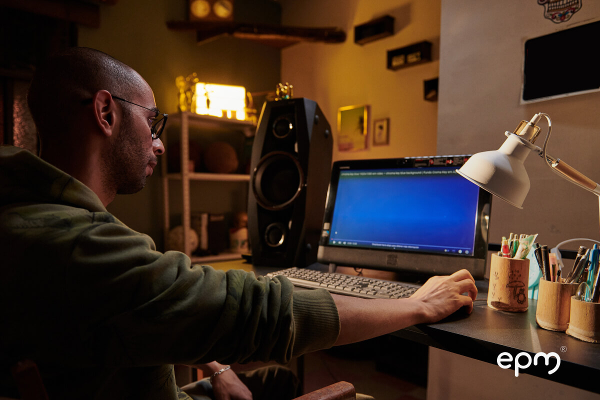 Hombre usando un computador de escritorio en una habitación con iluminación cálida, lámpara encendida, parlante grande y útiles sobre el escritorio