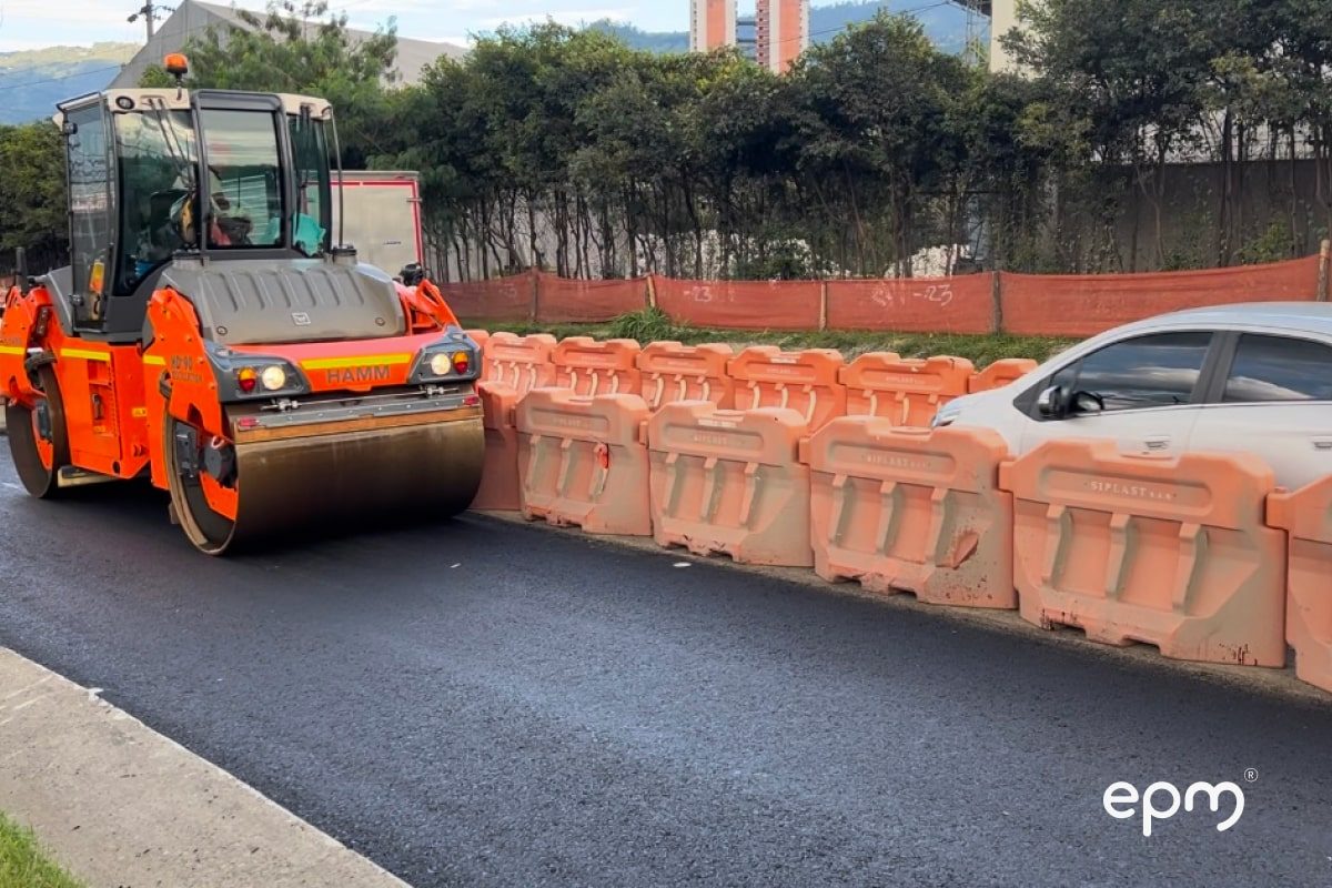 Máquina compactadora asfáltica trabajando en la pavimentación de una vía, con barreras de seguridad naranjas y un vehículo circulando al lado.