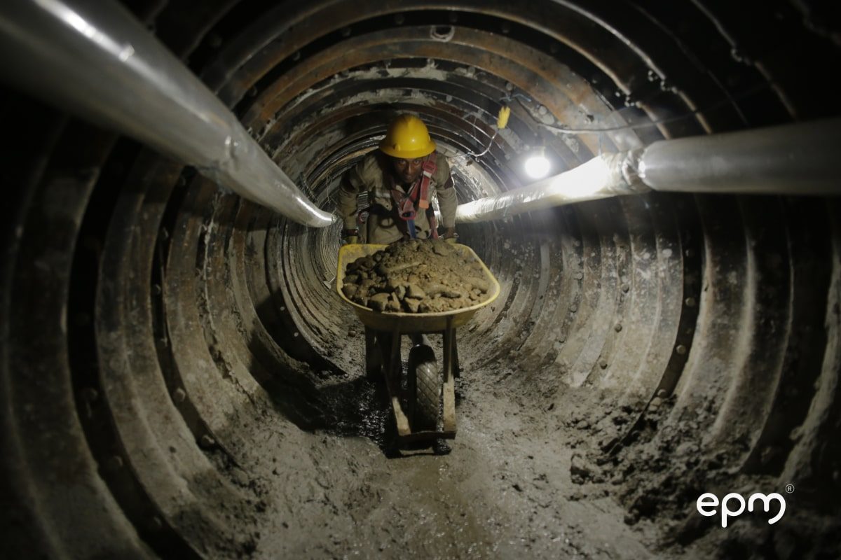 Trabajador con casco y arnés transporta escombros en una carretilla dentro de un túnel en construcción, iluminado con luces artificiales.