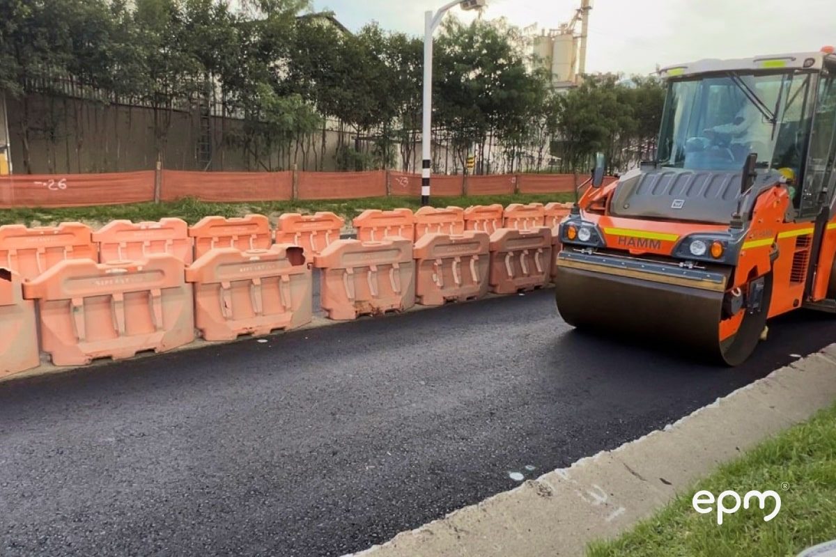 Máquina compactadora asfáltica trabajando en la pavimentación de una vía, con barreras de seguridad naranjas a los lados.
