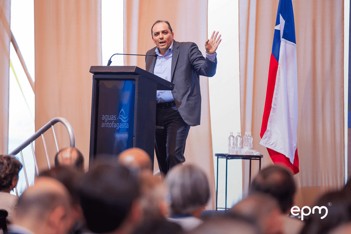 Fotografía de representante del Grupo EPM dando un discurso en el evento de Aguas Antofagasta, con público asistente y la bandera de Chile al fondo.