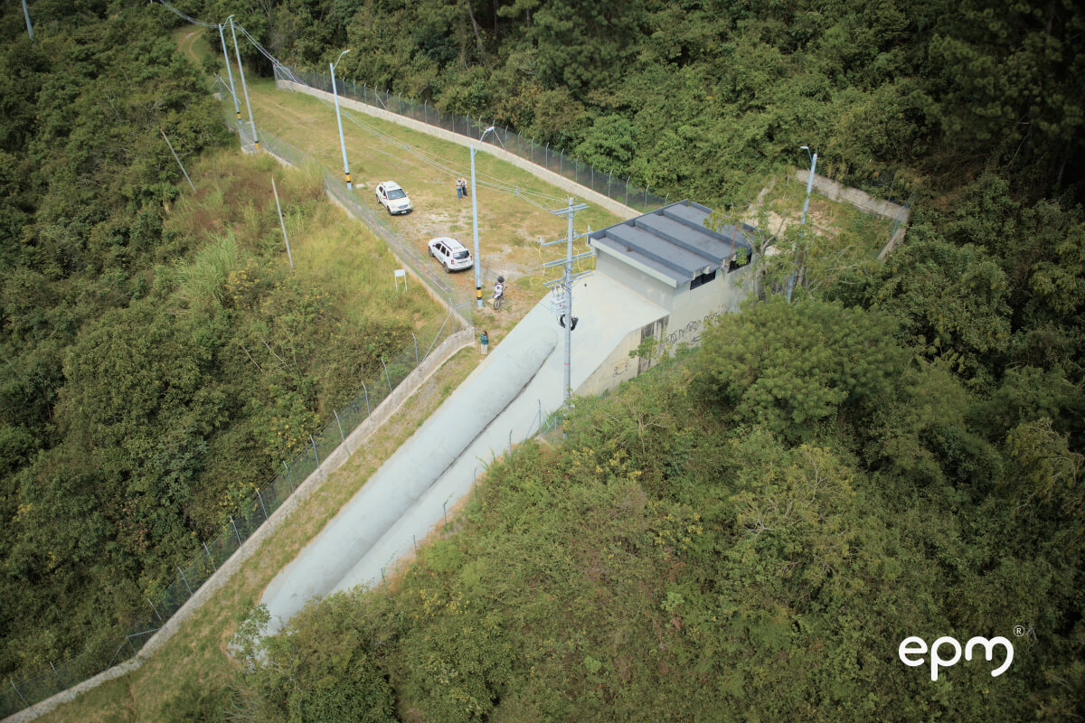 imagen de una planta de tratamiento de agua rodeada de vegetación, con una carretera que conduce a la planta y varios vehículos estacionados cerca. La imagen está etiquetada como "epm".