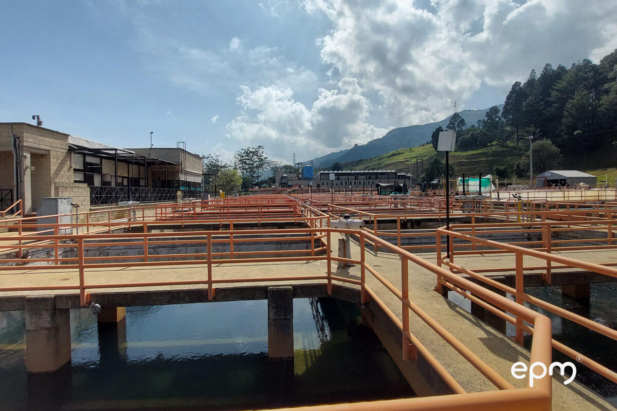 Imagen de una planta de tratamiento de agua con múltiples tanques y pasarelas, con montañas y un cielo parcialmente nublado de fondo. La imagen está etiquetada como "epm".