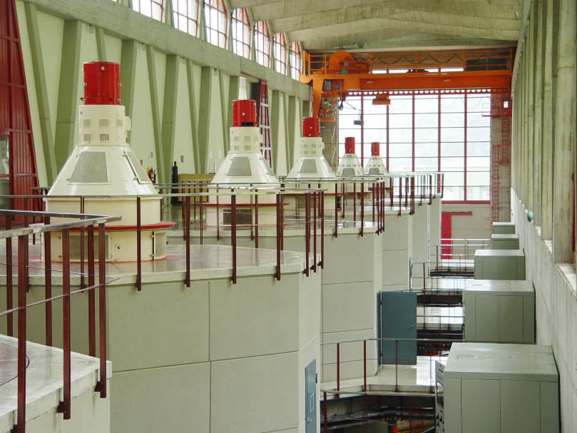 Interior de una planta hidroeléctrica con varias turbinas alineadas, protegidas por barandas rojas y estructuras blancas con cabezales rojos.