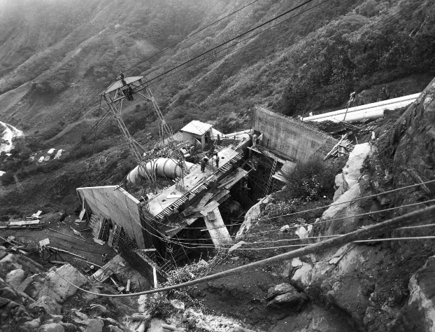 Fotografía histórica en blanco y negro de una obra de infraestructura hidroeléctrica en una zona montañosa, con estructuras metálicas, tuberías de gran tamaño y cables suspendidos.