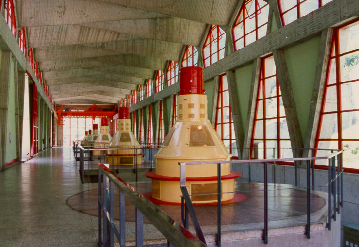 Sala de máquinas de una central hidroeléctrica con turbinas de color amarillo y rojo alineadas bajo una estructura con grandes ventanales y techo de concreto curvado.