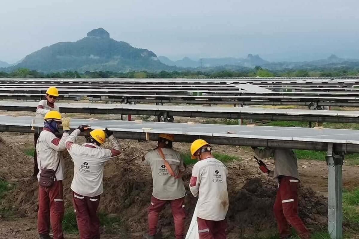 Fotografía de un grupo de trabajadores con cascos y uniformes instala la estructura de soporte para los paneles solares en una zona rural con montañas al fondo.