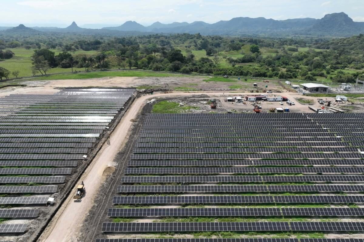Fotografía de vista aérea de un extenso parque solar con filas de paneles solares organizados, rodeado de vegetación y montañas en el horizonte.