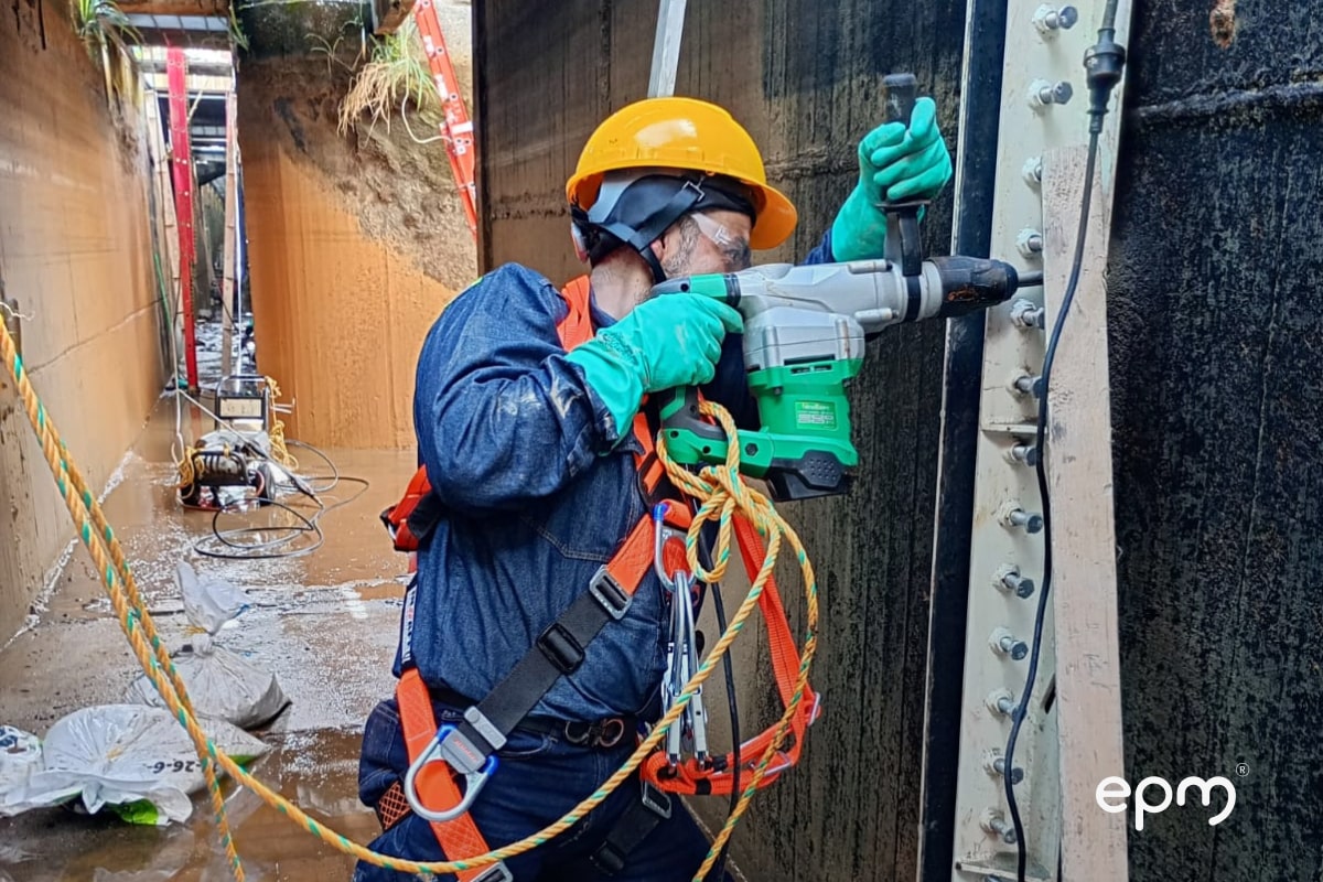 Trabajador con casco, gafas y arnés utiliza un taladro eléctrico para perforar una estructura metálica en un canal de concreto.