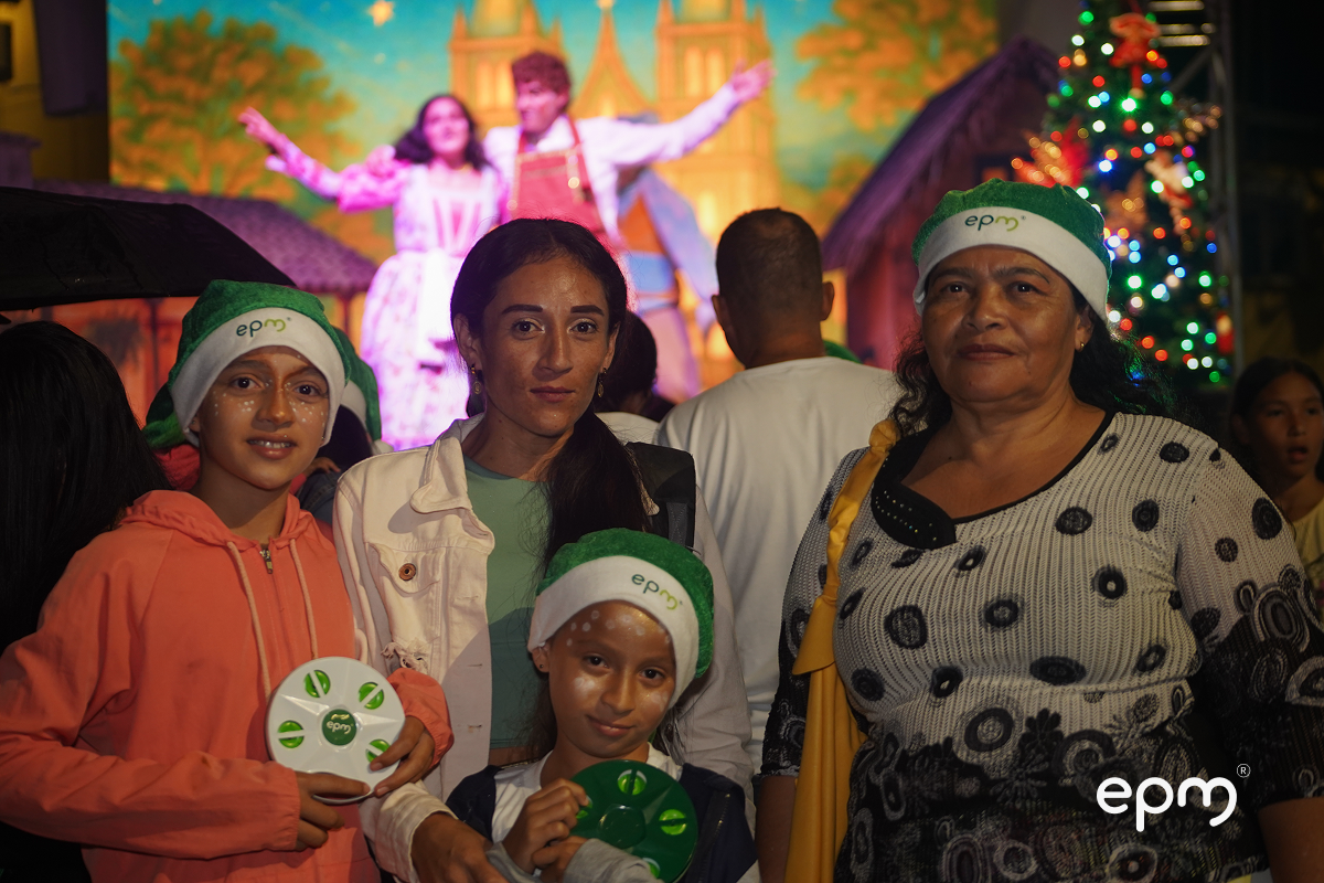 Mujeres acompañadas de niños con gorros de navidad. De fondo se observan actores de obra de teatro y árbol de navidad alumbrando