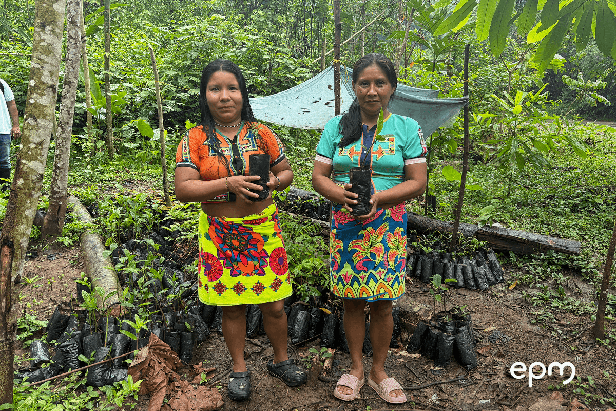 Fotografía de dos mujeres indígenas posan en un vivero forestal en medio de una zona boscosa, sosteniendo bolsas con plántula