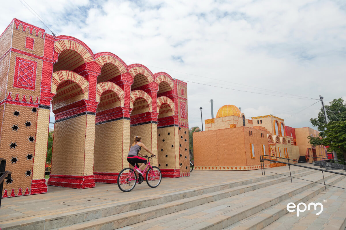 Mujer montando bicicleta frente a una colorida estructura decorativa con arcos de inspiración arquitectónica árabe. Escena parte de los alumbrados navideños de Medellín.