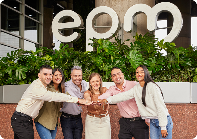 Grupo de seis personas sonrientes uniendo sus manos frente al edificio de EPM, con el logo grande de la empresa visible detrás y plantas decorativas alrededor