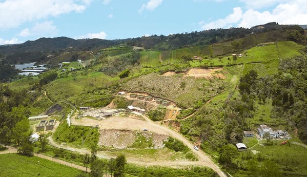 Vista aérea de una zona montañosa con vegetación verde, en la que se observan terrenos de cultivo y áreas de tierra removida, con algunas construcciones visibles en el fondo.