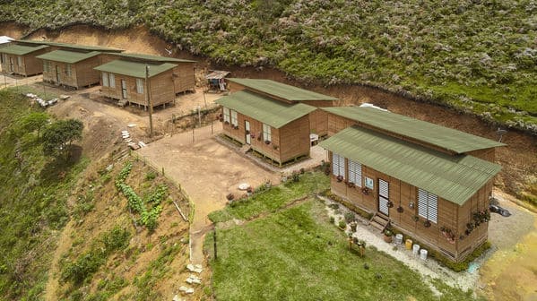 La imagen muestra un conjunto de casas de madera con techos de color verde. Están ubicadas en un terreno montañoso, rodeadas de vegetación.