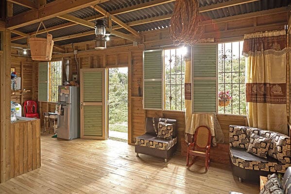 Interior de una casa de madera con muebles sencillos, luz natural entrando a través de ventanas abiertas, y una vista al exterior.