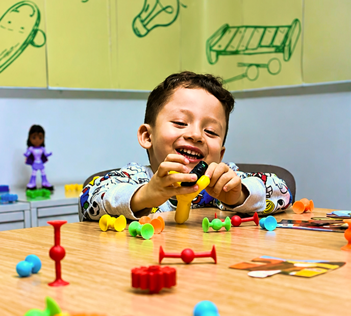 Niño sonriente jugando con piezas de colores sobre una mesa, en un espacio creativo con ilustraciones infantiles al fondo.