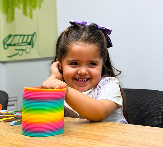 Niña sonriendo mientras juega con un resorte multicolor sobre una mesa, en un entorno lúdico y creativo.