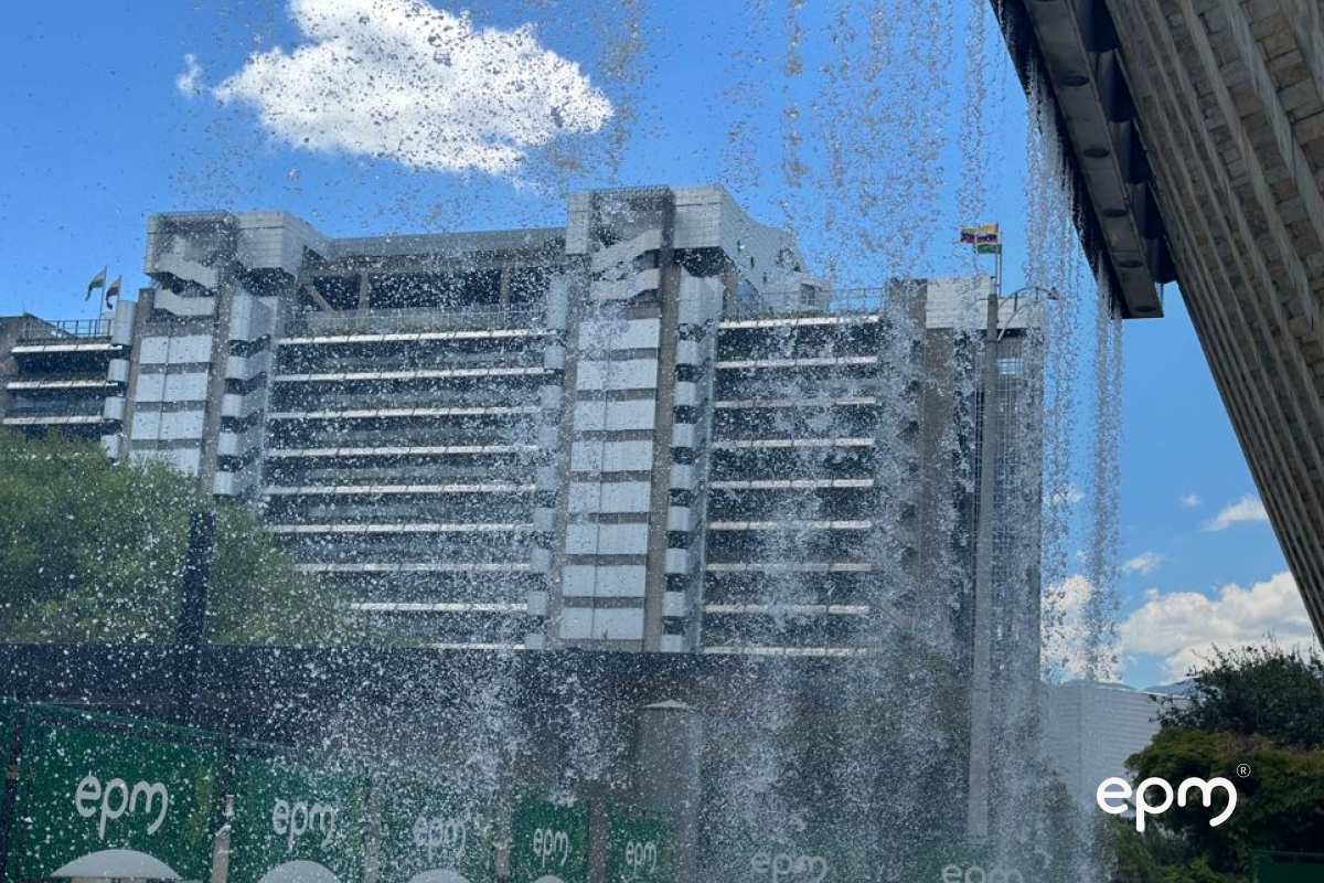 Se ve un edificio moderno a través del agua que cae con un cielo azul y nubes en el fondo; los logotipos "epm" son visibles en primer plano.