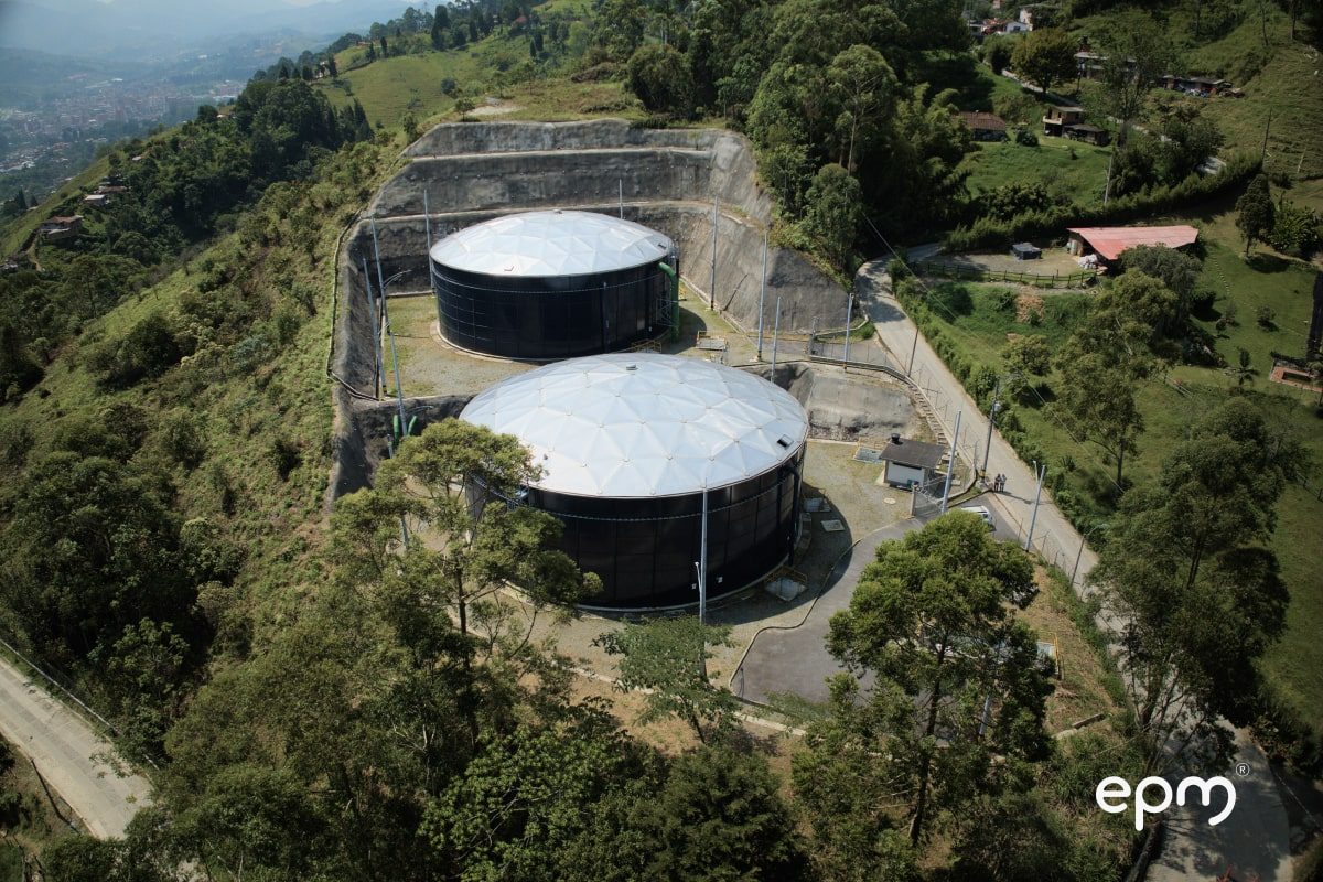 Photograph of an aerial view of two large water storage tanks with geometric roofs in a mountainous and green area.