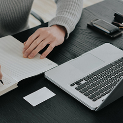 Persona escribiendo en una libreta junto a un computador portátil sobre una mesa de trabajo.