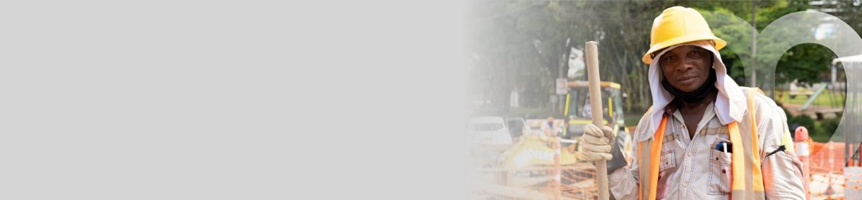 Trabajador de EPM con casco amarillo y chaleco naranja, sosteniendo una pala en un sitio de obra al aire libre.