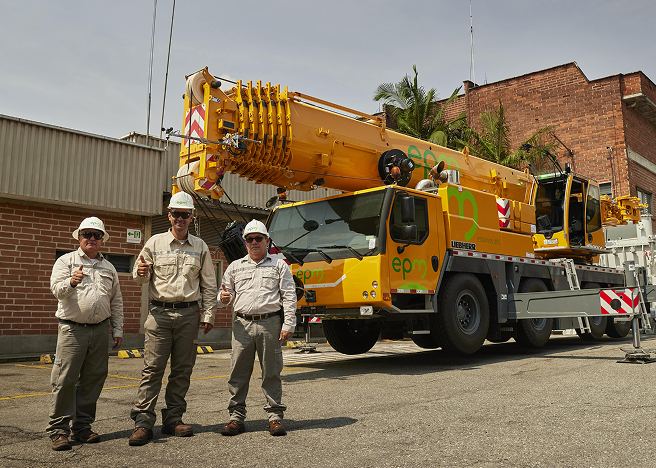 Tres trabajadores con cascos y uniformes de seguridad posan frente a una grúa amarilla de gran tamaño con el logo de EPM